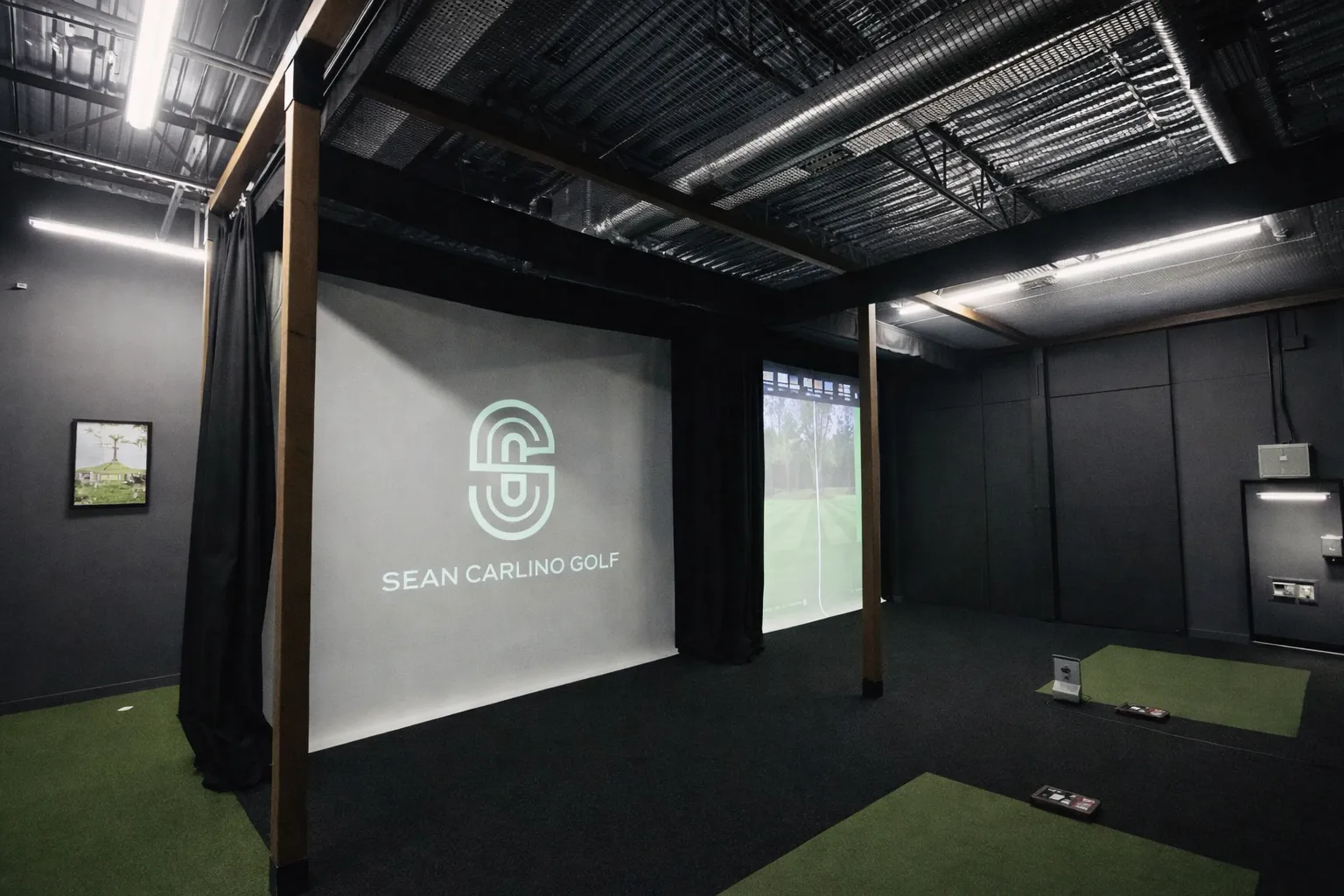 An indoor golf simulator studio with a "Sean Carlino Golf" logo projected onto a large white hitting screen, flanked by black curtains and multiple hitting mats.