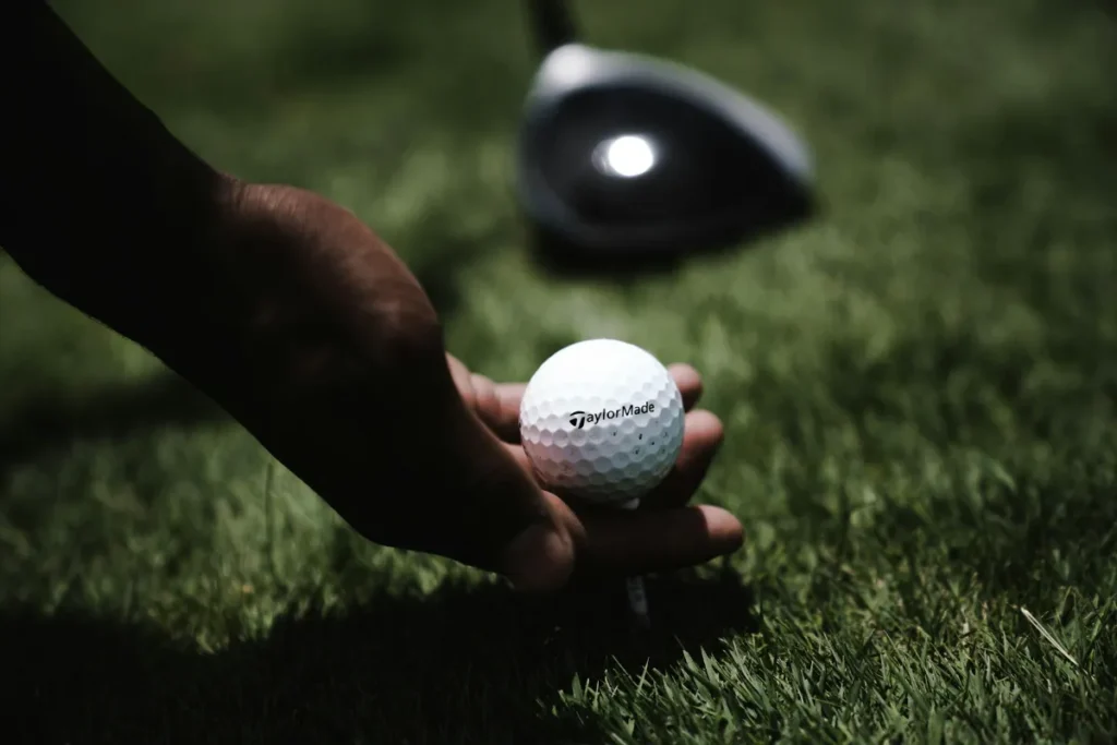 A hand carefully placing a white golf ball on a tee with a driver club resting in the background.
