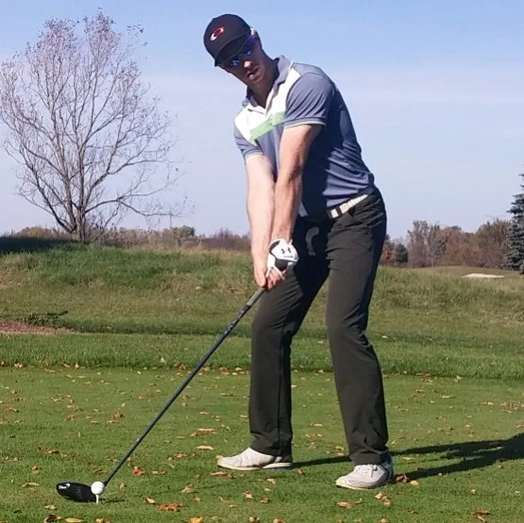A golfer in a blue polo and black cap addressing the ball with a driver on a green tee box under a clear blue sky.