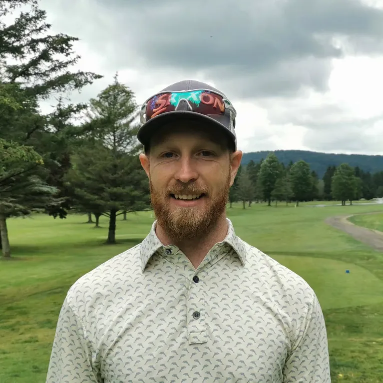 A smiling man with a red beard wearing a patterned polo shirt and a grey baseball cap with sunglasses on top, standing in front of a green golf course.