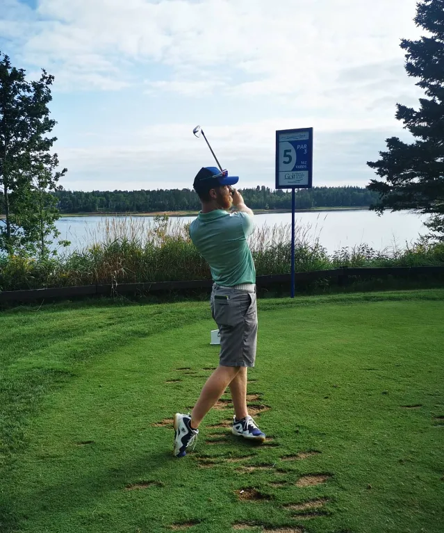 A man with a beard wearing a green polo shirt and a blue cap completing a golf swing on a tee box overlooking a lake, with a sign indicating Hole 5, Par 3 in the background.