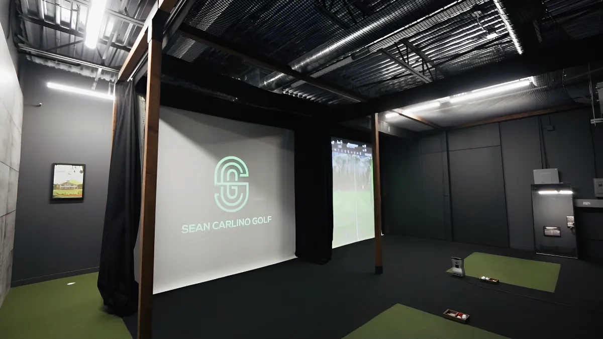 An indoor golf simulator studio with a sleek, modern design, featuring a large projection screen displaying a circular logo, green hitting mats, and black walls under an industrial-style ceiling.