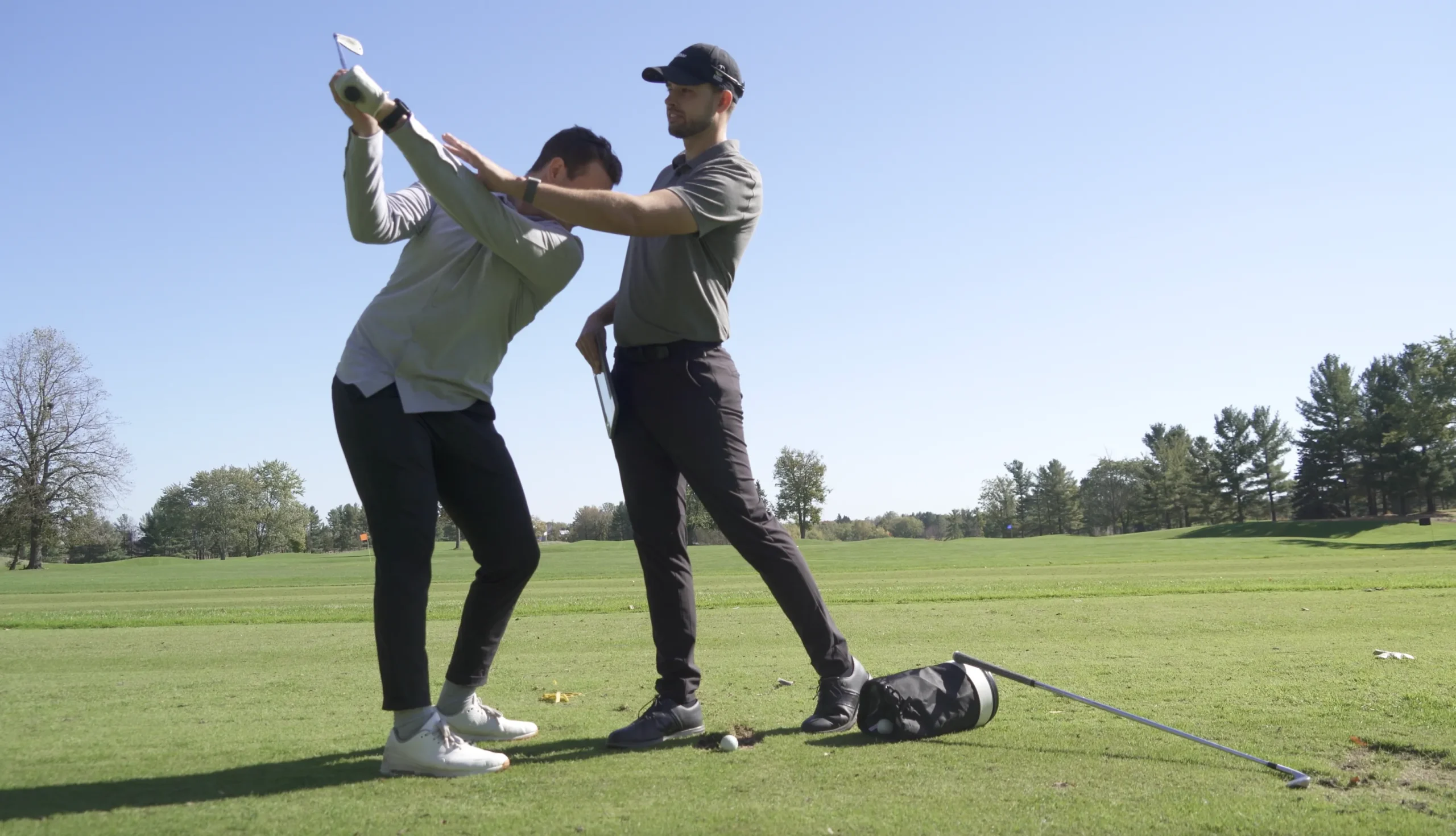 A professional golf instructor in a dark polo and cap stands behind a student on a bright, sunny golf course, physically adjusting the student's shoulder alignment during their backswing.
