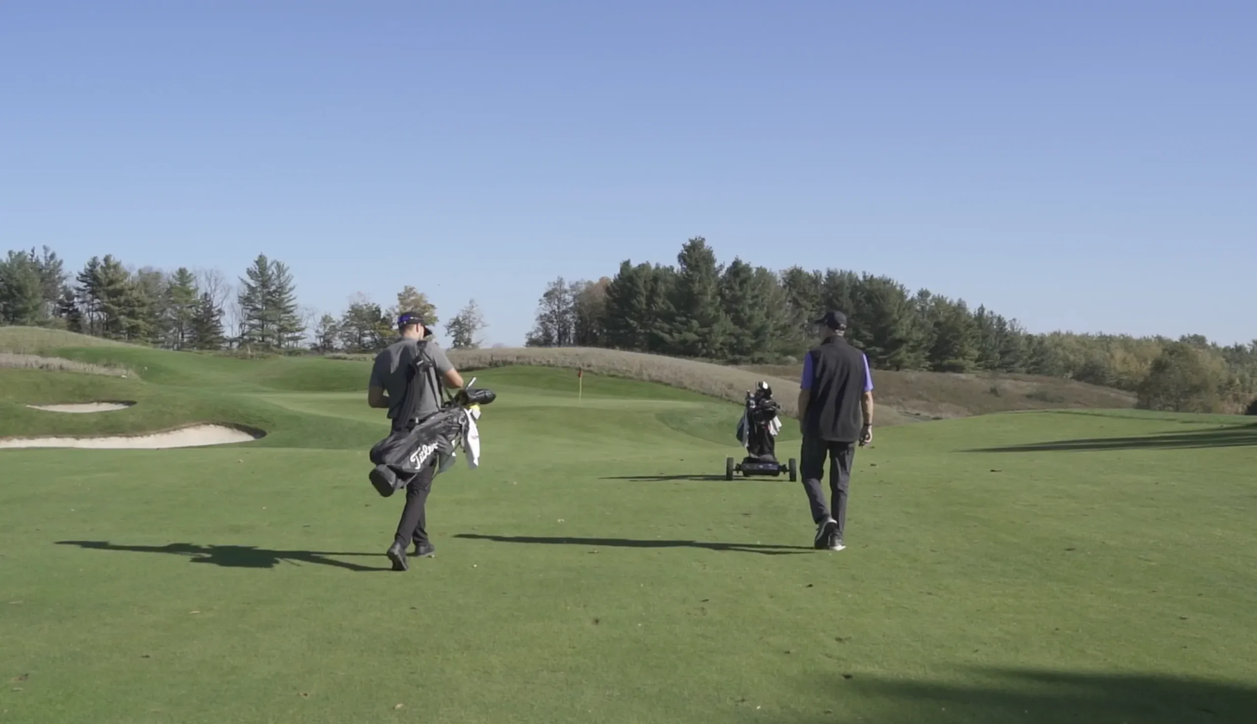 Two golfers carrying their bags while walking across a wide, green fairway under a clear blue sky.