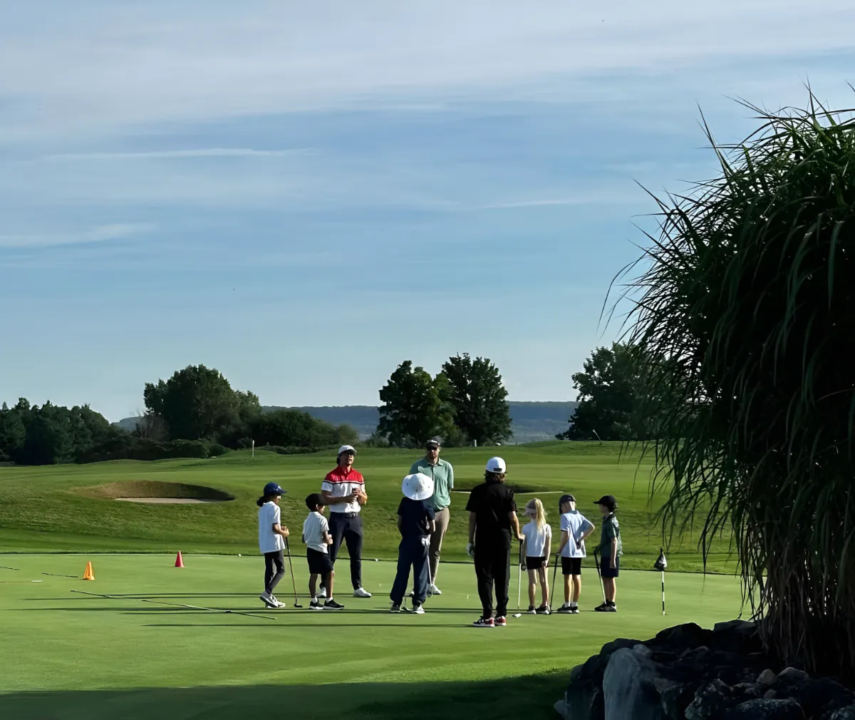 A group of young students and an instructor stand in a circle on a sunlit putting green for a lesson.