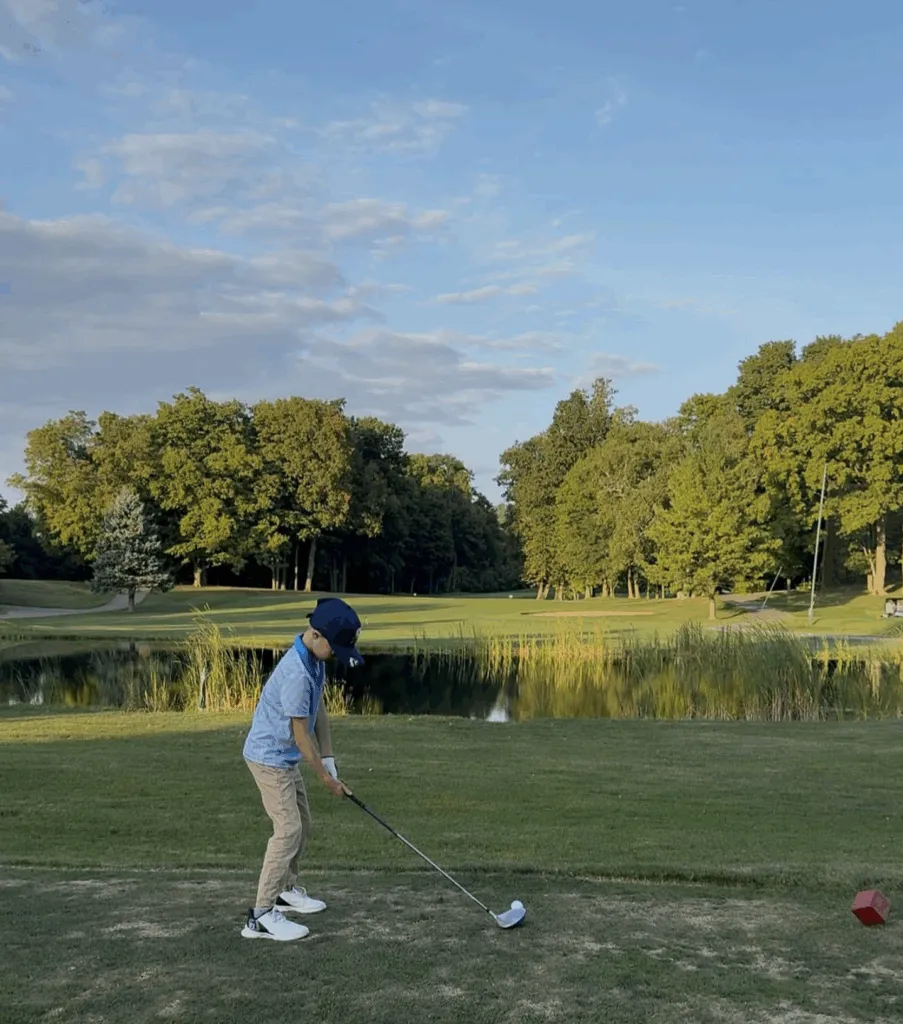 A young boy in a blue shirt and hat practicing his golf swing on a green fairway, with a small pond and a line of lush trees in the background under a clear sky.