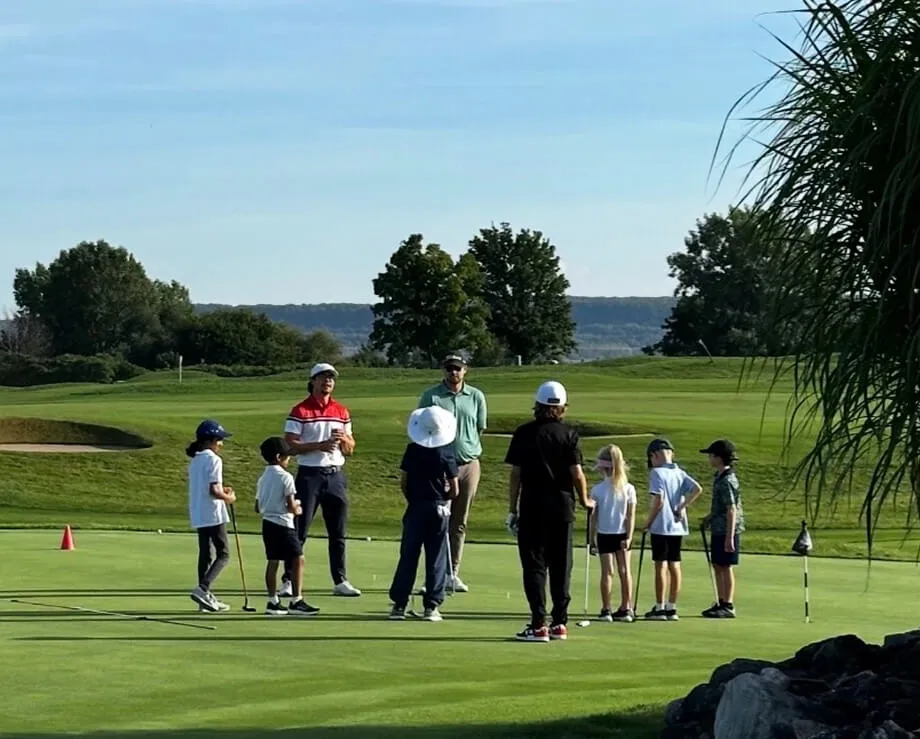 A group of junior golfers standing in a circle on a practice putting green. Two instructors are leading the session, with a scenic view of the golf course and a body of water in the distant background under a clear sky.