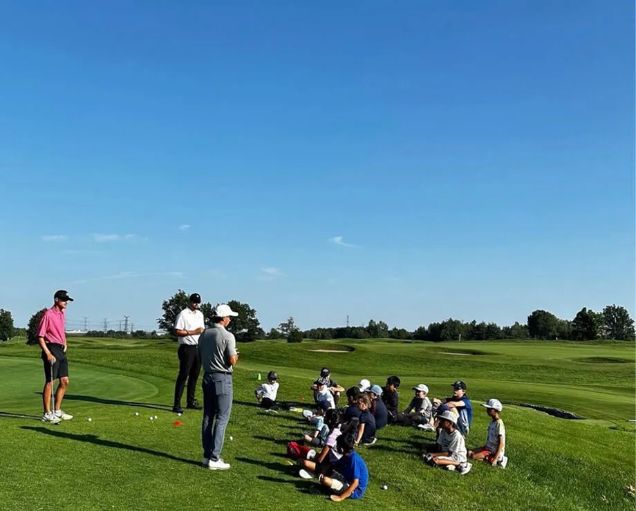 A wide shot of a junior golf clinic on a sunny day. A group of children are sitting on a grassy slope while two instructors stand and address the group. The scene is set on a vast, green golf course under a clear blue sky.