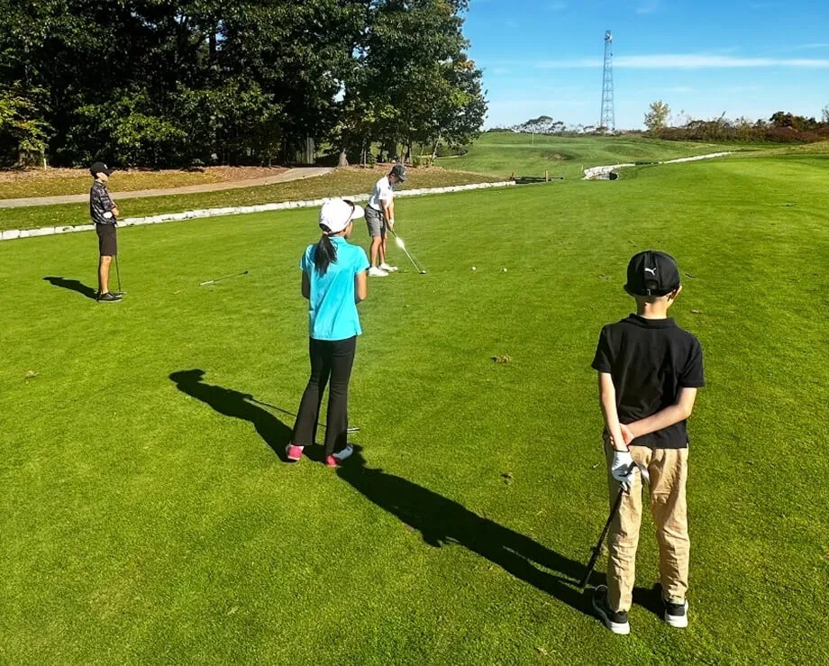 A group of junior golfers standing on a vibrant green fairway during an outdoor lesson. Several children are lined up on the grass under a clear blue sky, with a lush treeline in the background.