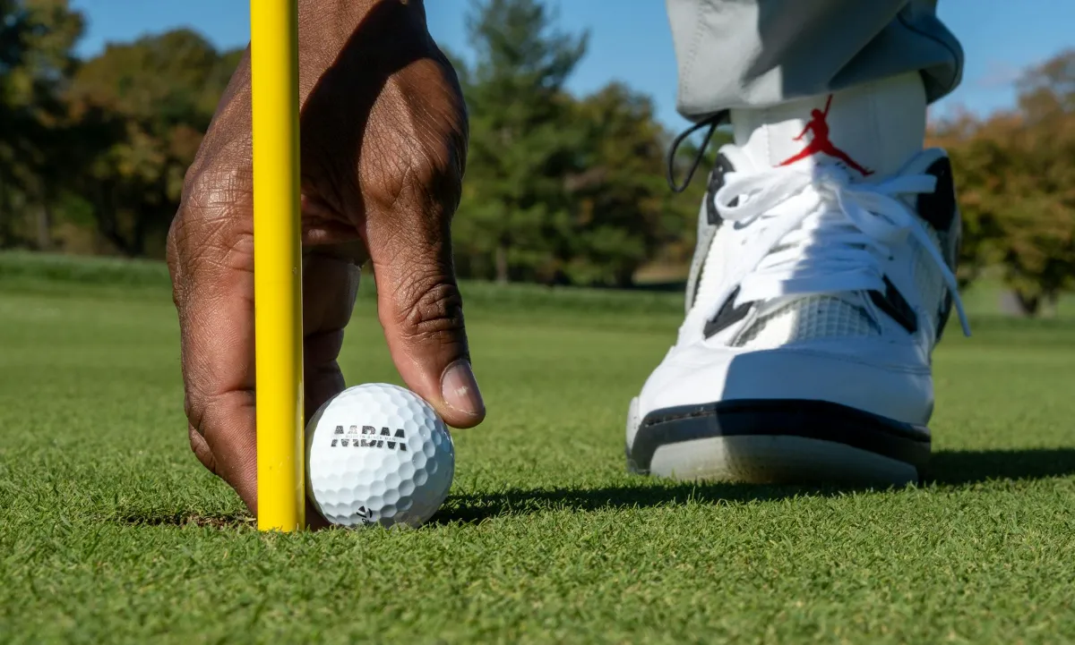 A hand places a golf ball into a hole next to a yellow flagstick on a green.