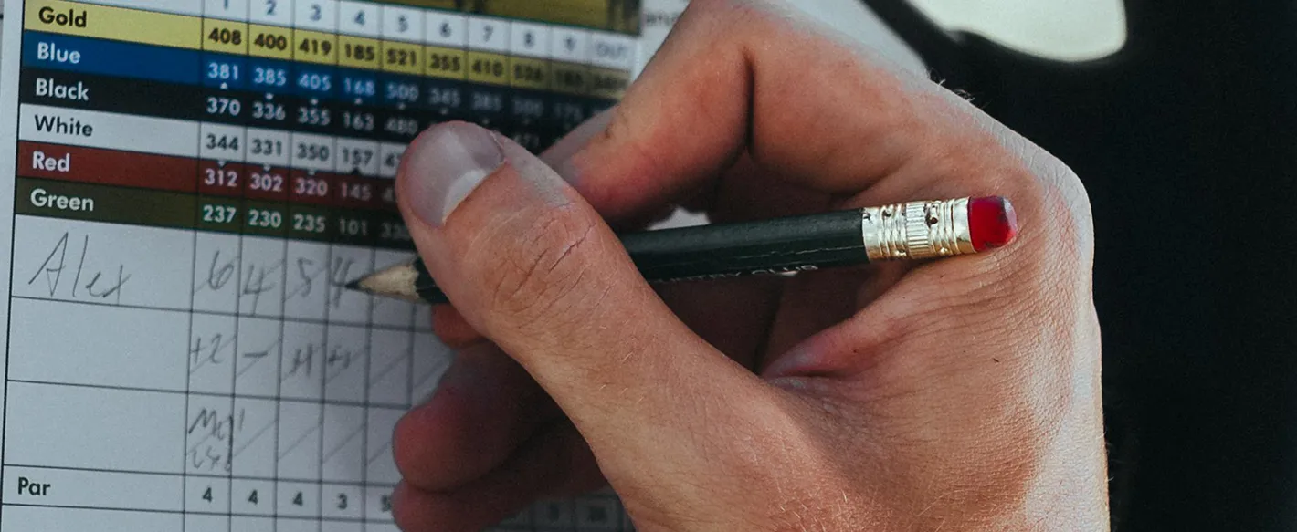 A close-up shot of a person's hand using a pencil to write scores on a colorful golf scorecard.