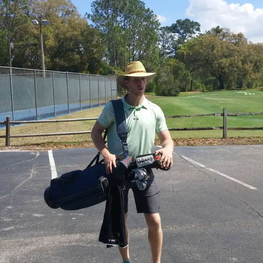 A male golfer in a straw hat carrying a black golf bag across a paved area toward a sunny green course.