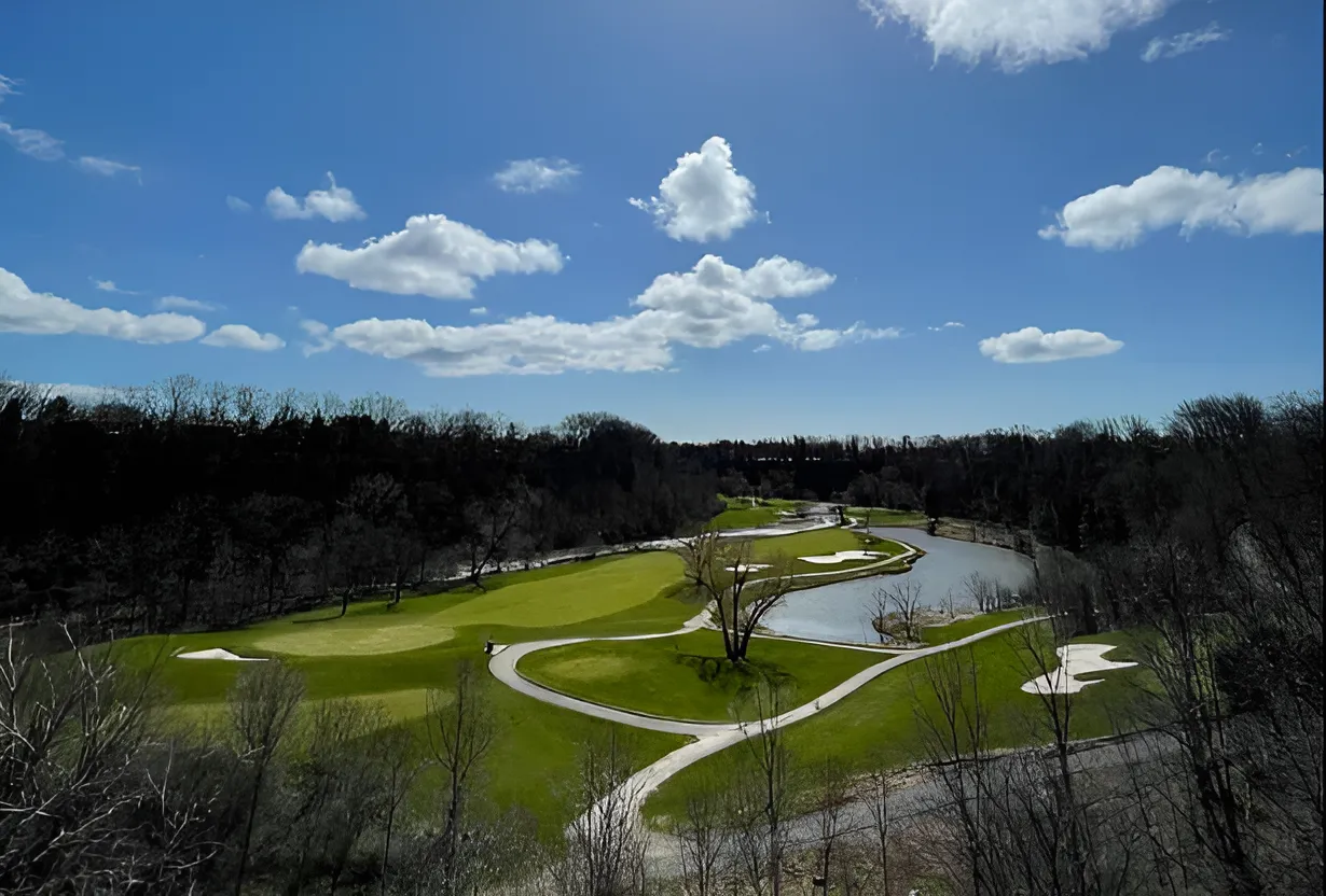 An elevated view of a vibrant green golf course nestled within a dense forest, featuring a winding white cart path, a large water hazard, and bright white sand bunkers under a blue sky with scattered clouds.