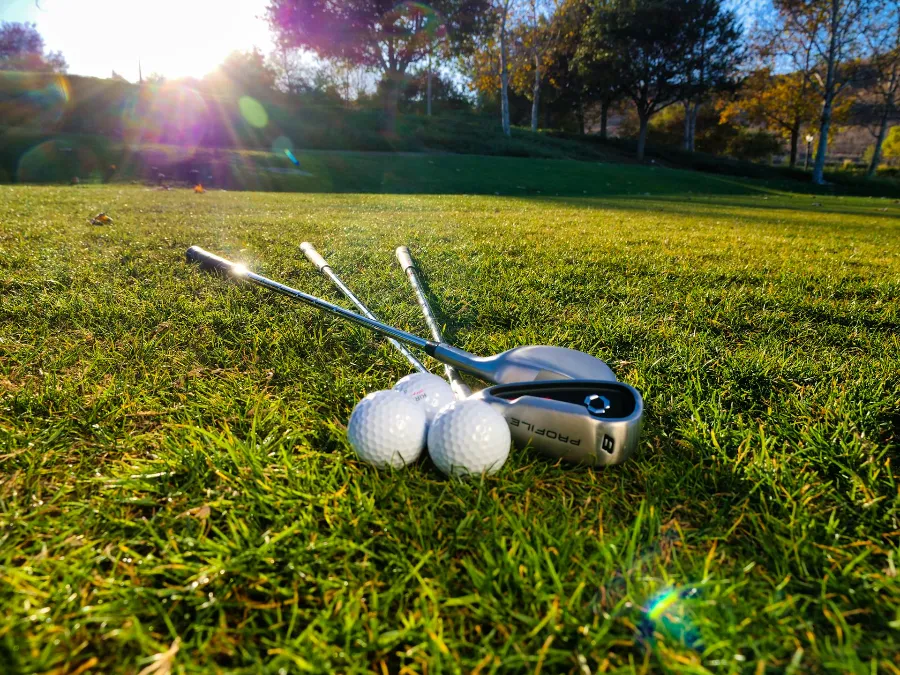 A close-up, low-angle shot of three white golf balls nestled in the grass next to a golf club. The scene is illuminated by a warm sunset with a bright lens flare, and a lush treeline is visible in the soft-focus background.