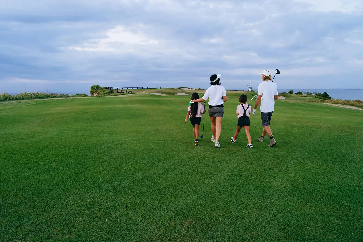 A family of four walks away from the camera across a wide, green golf course toward the horizon under a cloudy sky.