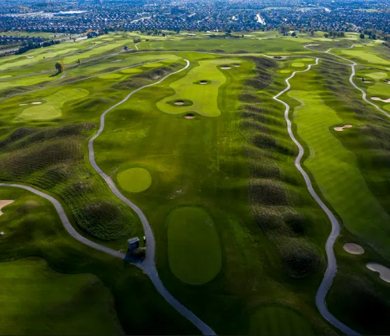 An aerial, top-down view of a golf course featuring distinct undulating grassy mounds, winding paved cart paths, and several greens under soft lighting that emphasizes the rolling terrain.