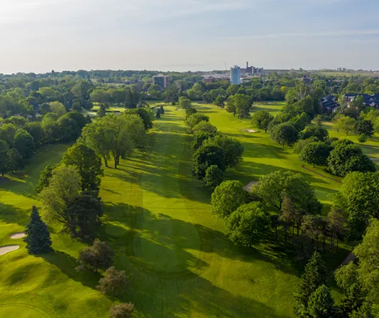 An aerial view of a vibrant green golf course fairway lined with mature trees, showing long shadows and a distant building on the horizon.