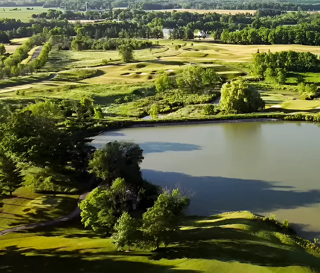 An aerial view of a golf course featuring a large pond in the foreground, surrounded by vibrant green fairways and groves of leafy trees under bright, sunny conditions.