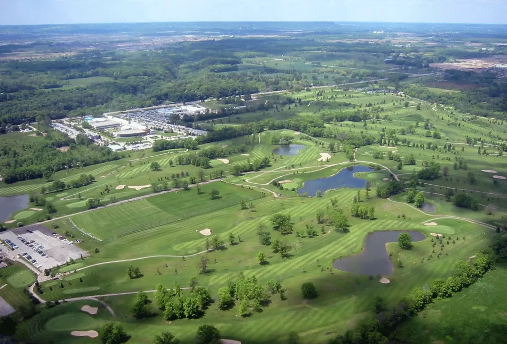 An expansive aerial view of a golf course featuring undulating green fairways, several small ponds, and sand bunkers, with a large clubhouse and parking area visible in the background under a clear sky.